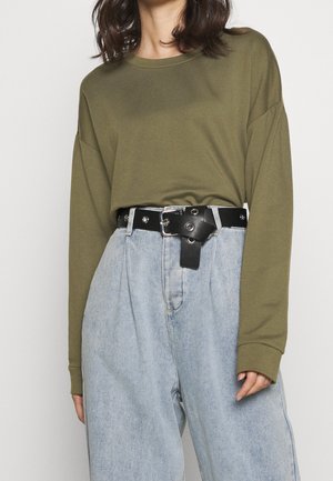 Olivgroen top met lange mouwen gecombineerd met een lichtblauwe, hooggesneden spijkerbroek en een zwarte tailleband met zilverkleurige accenten.