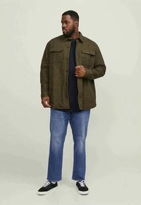 Homem com uma camisa verde oliva de botões sobre uma t-shirt preta, jeans azuis e sneakers pretos, de pé contra um fundo claro e liso.