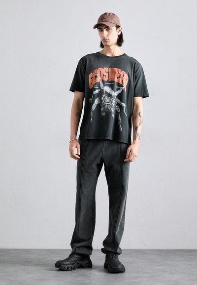 T-shirt graphique noir avec design de toile d'araignée et texte orange, assorti à un pantalon gris foncé avec coutures latérales blanches et des baskets noires imposantes.