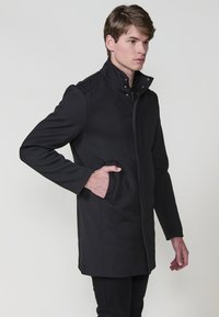 Abrigo de lana negro con un alto cuello, cremallera frontal completa y bolsillos laterales. Diseño elegante con una tela texturizada y sutiles detalles de costura.