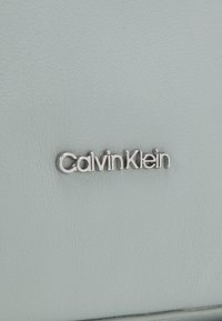 Logo argenté Calvin Klein sur une surface en cuir texturé gris clair, centré et clairement embossé.