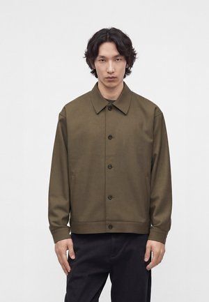 Homme portant une veste boutonnée vert olive avec col et un pantalon noir, debout devant un fond clair uni.