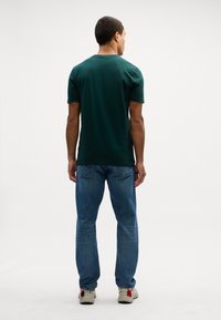 Camiseta de algodão verde escuro com mangas curtas, gola redonda, ajuste standard, combinada com jeans de ganga azuis e ténis de cor clara.