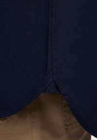 A camisa de algodão azul-marinho apresenta uma bainha curva com costuras contrastantes, combinada com calças texturizadas castanho-claro parcialmente visíveis abaixo.