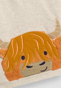 Ljust grått tyg med en broderad höglandsko i brunt och orange, med en rufsig man och två böjda horn, mot en texturerad bakgrund.