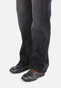 Svarte denim jeans kombinert med to forskjellige stilte sneakers: en med svarte lisser og en med brune lisser, med teksturerte overflater og mørke nyanser.