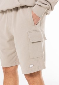 Beige kravas šorti, izgatavoti no mīkstas kokvilnas. Iekļauj sānu kabatu ar vāku un logotipa detaļu. Elastīgs jostasvīrs komfortam.