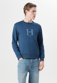 Felpa in cotone blu navy con maniche lunghe, collo a giro e grafica frontale con una "H" e il testo "HACKETT". Polsini e orlo a coste.