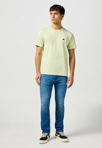 Ljusgrön kortärmad t-shirt med rund hals, matchad med blå jeans och svarta sneakers med cremefärgade accenter. Litet märke på t-shirten.