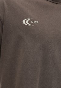 Καφέ ύφασμα με μικρό λευκό λογότυπο ARKK που απεικονίζει δύο κυρτές γραμμές στην πάνω περιοχή του στήθους.