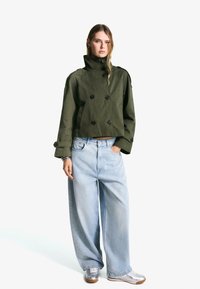 Avskuren olivgrön dubbelt knäppt jacka med stora svarta knappar, parat med lösa ljusblå denimjeans och silversneakers.