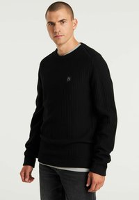 Maglione nero in maglia waffle con scollatura a girocollo a coste, caratterizzato da un piccolo logo sul petto. Maniche lunghe e vestibilità ampia.