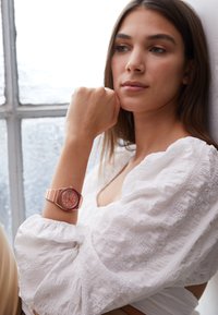 Montre rose avec un bracelet rayé, un cadran rond et des accents argentés. Un chemisier blanc texturé et un éclairage doux créent une ambiance lumineuse.