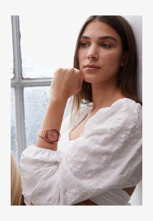 Roze horloge met gestreepte band, ronde wijzerplaat en zilveren accenten. Een getextureerde witte blouse en zachte verlichting creëren een heldere setting.