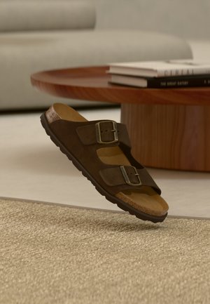 Sandalo in suede marrone con due cinturini fibbia, suola in sughero, suola nera, vicino a un tavolo rotondo in legno e tappeto beige in un ambiente soggiorno.