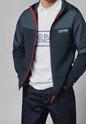 Mann trägt eine marineblaue und dunkelgraue Red Bull Racing Reißverschlussjacke über einem weißen Poloshirt mit Logo und dunkelblaue Jeans, Hände in den Taschen.