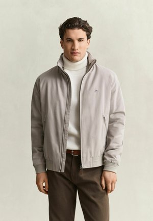 Junger Mann trägt eine hellgraue Jacke mit Reißverschluss über einem weißen Rollkragenpullover und dunkelbraunen Hosen, steht vor einem schlichten Hintergrund.
