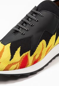 Zapatilla negra con un patrón de llamas en amarillo y rojo, con detalles de tejido texturizado y cuero liso. Incluye cordones a rayas en blanco y negro.