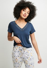 T-shirt blu navy a scollo a V con rifiniture in pizzo, abbinato a pantaloni bianchi con un motivo floreale in giallo, blu e viola. Tessuto morbido, vestibilità rilassata.