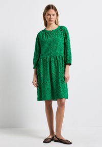 Abito verde a lunghezza ginocchio realizzato in tessuto testurizzato, con motivi a taglio, scollatura rotonda e maniche tre quarti. Scarpe flat nere.