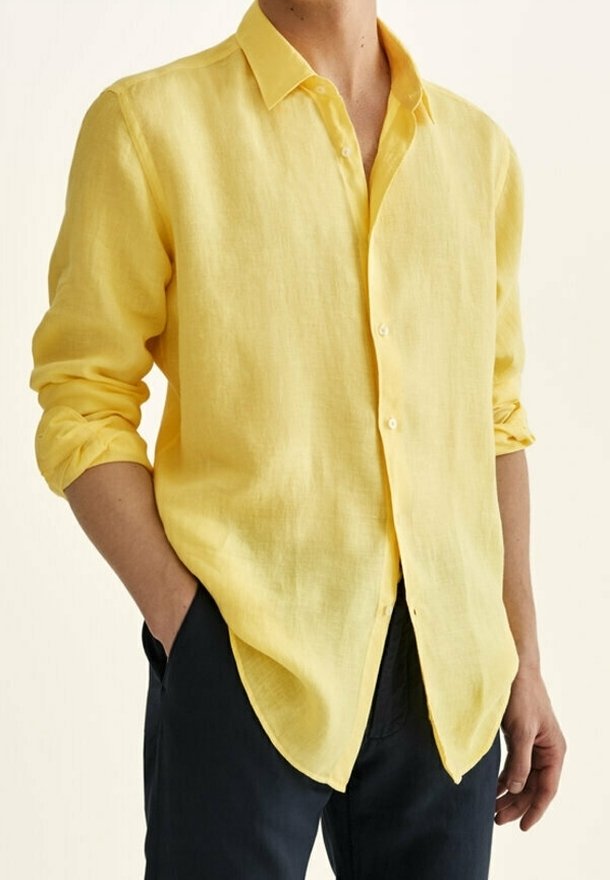 Personne portant une chemise jaune à manches longues avec les manches retroussées et un pantalon foncé, les mains dans les poches, debout devant un fond uni.