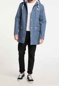 Blå vattentät jacka med huva, som har flera fickor, knapppdetaljer och dragkedjestängning. Bärs över en vit skjorta.