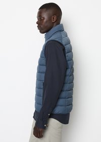 Blauer Steppweste mit hohem Kragen, gepolstertem Design und Seitentaschen, getragen über einem dunklen Hemd. Glatte Textur und elegante Silhouette.