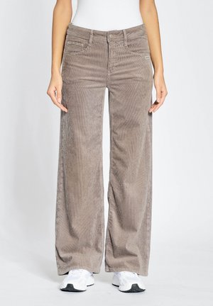 Osoba ubrana w beżowe szerokie spodnie sztruksowe i białe trampki, stojąca na jednolitym jasnym tle.