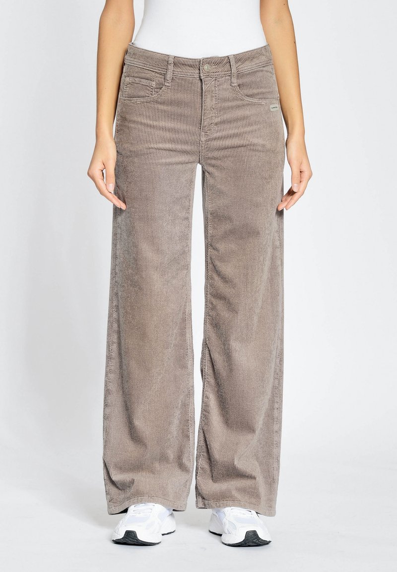 Henkilö, joka pukeutunut beigeihin leveälahkeisiin vakosamettihousuihin ja valkoisiin tennareihin, seisoo yksinkertaisen vaalean taustan edessä.