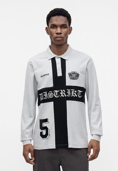 Homme portant un polo gris à manches longues avec un motif de croix noire, le texte "DISTRIKT", le numéro 5 et un emblème de blason sur la poitrine.