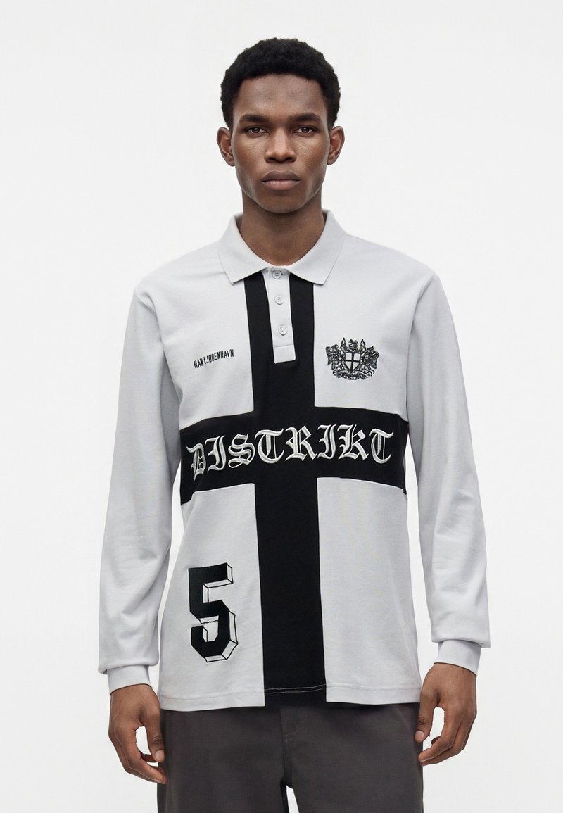 Homme portant un polo gris à manches longues avec un motif de croix noire, le texte "DISTRIKT", le numéro 5 et un emblème de blason sur la poitrine.