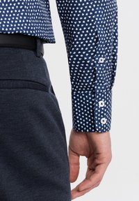 Camicia blu navy con bottoni e motivi a foglia bianchi, in tessuto di cotone, con colletto aderente. Pantaloni scuri e strutturati visibili in vita.