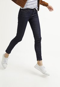 Vaqueros ajustados azul oscuro con textura suave, que presentan un diseño de cinco bolsillos y detalles de costura sutiles, combinados con zapatillas blancas.