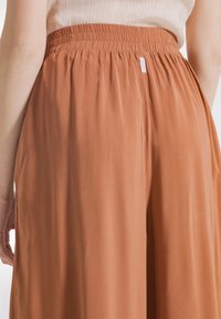 Terrakotta bukser med vide ben og elastisk talje; glat tekstur; diskret mærketag på bagsiden.
