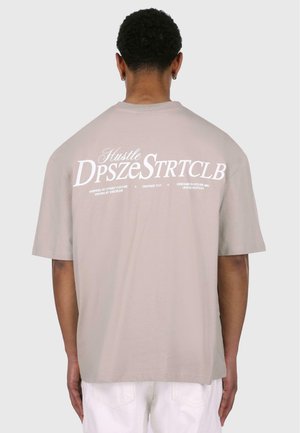 T-shirt oversize beige clair à manches courtes, avec du texte blanc et un design graphique au dos. Texture en coton lisse.