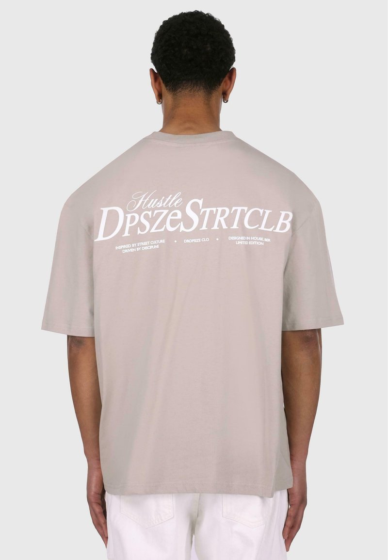 Lys beige oversized t-shirt med korte ærmer, prydet med hvid tekst og grafisk design på ryggen. Glat bomuldsstruktur.