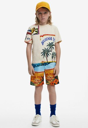Camiseta beige con gráficos coloridos, que incluyen palmeras y texto; combinada con pantalones cortos de patrón vibrante con motivos tropicales. Gorra amarilla.