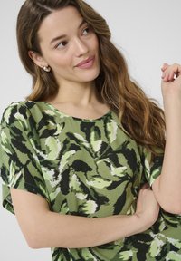 Lühikeste varrukatega tops, millel on roheline ja must lehtede print, laia kaeluse ja tekstuuriga kangaga. Lihtne disain, vaba lõige.