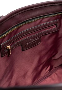 L'interno della borsa in pelle bordeaux presenta una tasca con zip e accessori in oro, una targhetta con logo in rilievo e una fodera in tessuto liscio a contrasto.
