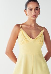 Mujer con el cabello recogido, lleva pendientes de aro dorados y un vestido amarillo sin mangas con tirantes finos, de pie contra un fondo blanco.