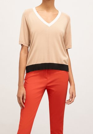 Persoon draagt een beige V-hals top met korte mouwen en een zwarte zoom en feloranje broek tegen een effen achtergrond.