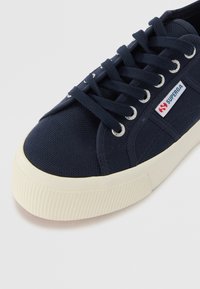 Námořnické plátěné tenisky s kulatou špičkou, bílou gumovou podrážkou, stříbrnými nýtky a modrou logovou nášivkou na boku. Tkaničky jsou pevně utažené.