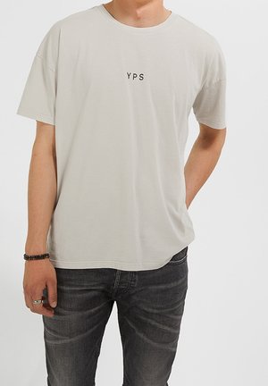 Jasnoszary bawełniany T-shirt z krótkimi rękawami, okrągłym dekoltem i czarnym napisem "YPS" na piersi, zestawiony z ciemnoszarymi dżinsami.