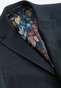 Blazer grigio scuro in tessuto testurizzato con fodera floreale in tonalità di blu, arancione e verde. Sono visibili i revers a scossa e le cuciture dettagliate.