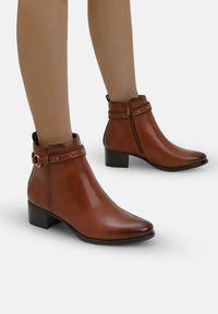 Botas de cuero marrón hasta el tobillo con punta redondeada, cremallera lateral, correa decorativa con tachuelas y tacón bajo grueso. Textura suave, diseño elegante.