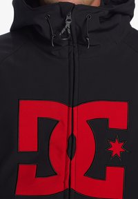 Čierna bundy na zips s kapucňou, vybavená veľkým červeným logom DC a akcentom hviezdy. Materiál sa zdá byť hladký s mierne textúrovaným povrchom.