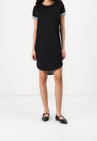 Svart kortärmad t-shirtklänning med grå ärmdetaljer, rund halsringning och en något längre bakre fåll, tillsammans med svarta platta skor.