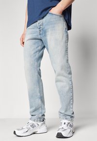 Jeans de denim azul claro con diseño de pierna recta, desvanecimiento sutil y bolsillos frontales, combinados con zapatillas deportivas blancas y grises.
