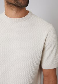 Jasno beżowy sweter z krótkim rękawem i fakturowanym splotem, z okrągłym dekoltem. Posiada subtelny wzór i prążkowane krawędzie.