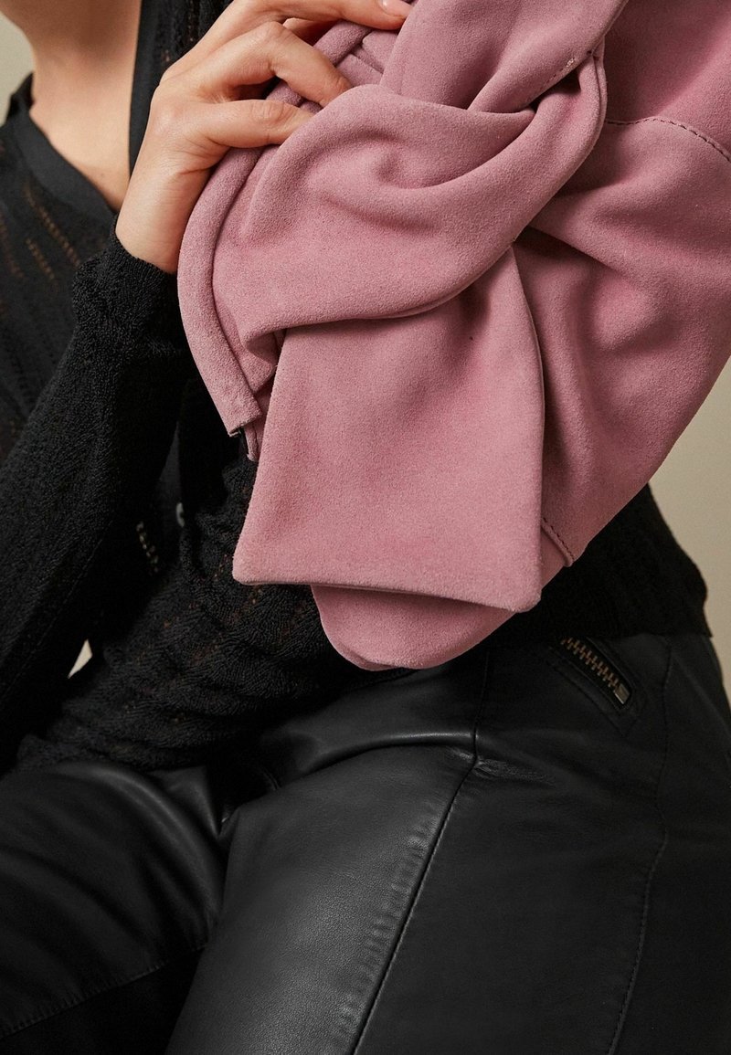 Tela rosa y suave caída en las manos sobre un suéter negro texturizado; pierna del modelo en cuero negro elegante. Detalle de cremallera notable en los pantalones.